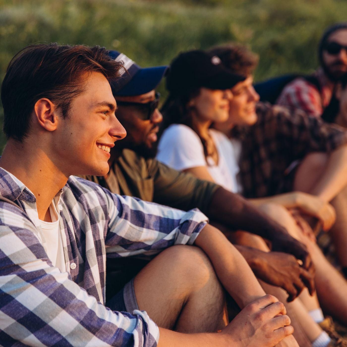 Five young adults sit outdoors in a row, smiling and enjoying a sunny day together.
