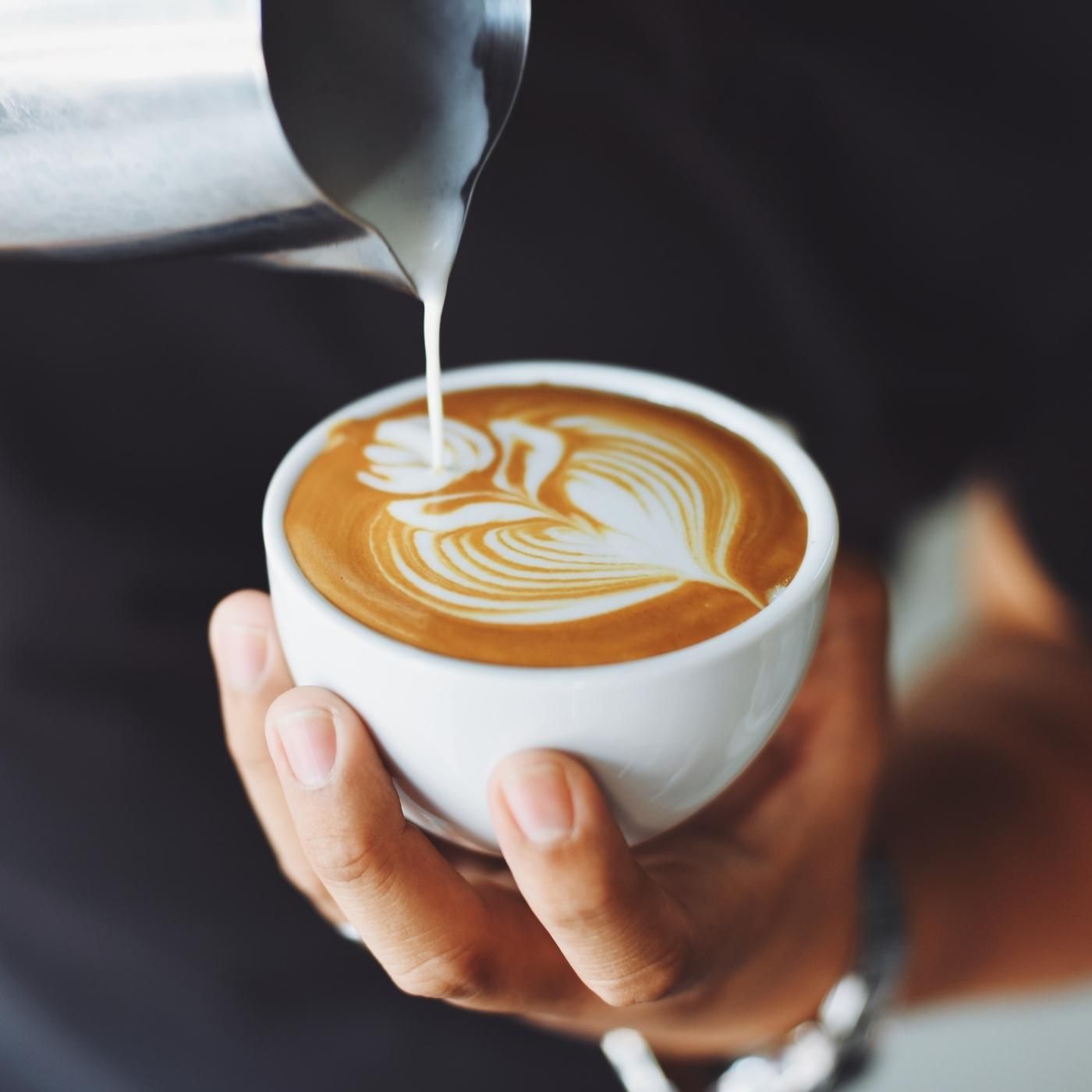 A hand holds a latte as milk is poured, creating intricate latte art on the coffee’s surface.