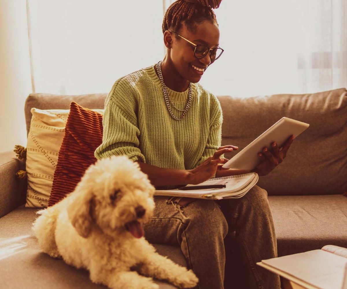 Woman sitting on a couch using a tablet, with a fluffy dog beside her and notebooks on her lap.